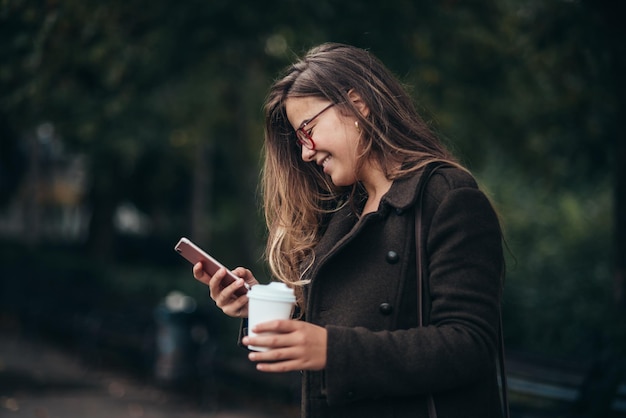 Piękna młoda kobieta za pomocą smartfona i pije kawę na wynos