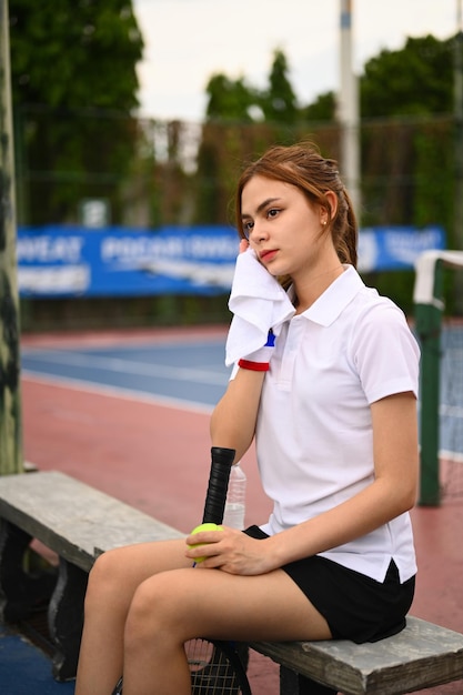 Piękna młoda lekkoatletka wycierająca pot ręcznikiem spoczywającym na ławce na korcie tenisowym po treningu