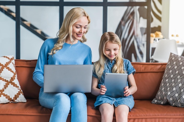 Piękna Młoda Mama I Jej Córka Używają Gadżetów I Uśmiechają Się Siedząc Na Kanapie W Domu