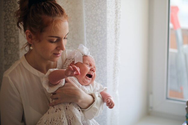 Piękna młoda mama i noworodek w jej ramionach stoją przy oknie