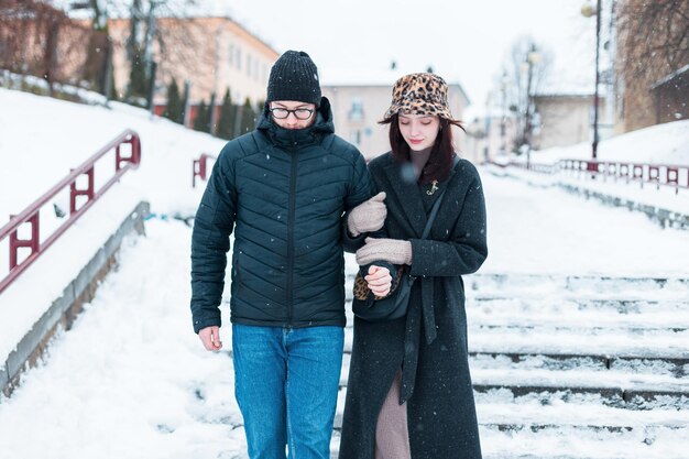 Piękna młoda modna para mężczyzna i kobieta w modnych zimowych ubraniach z kapeluszem i kurtką idą razem po mieście w zimowy dzień para kochanków schodzi po schodach