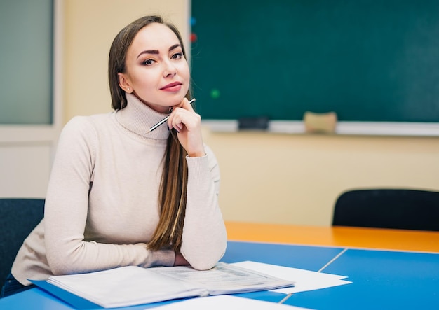 Piękna młoda nauczycielka z notatkami siedzi przy stole w klasie i uśmiecha się do kamery Tło tablicy