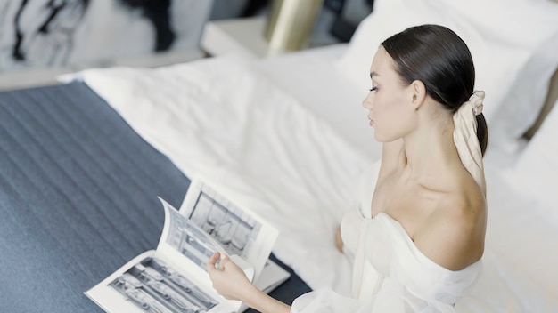 Zdjęcie piękna młoda panna młoda siedzi na łóżku i przewraca strony kobiety w białej sukience magazynu akcji