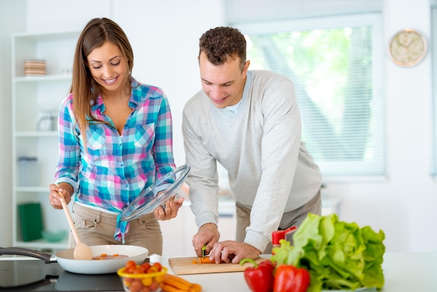 Piękna młoda para gotowanie zdrowego posiłku w domowej kuchni. Młody mężczyzna kroi marchewkę na desce kuchennej, a kobieta sprawdza posiłek.