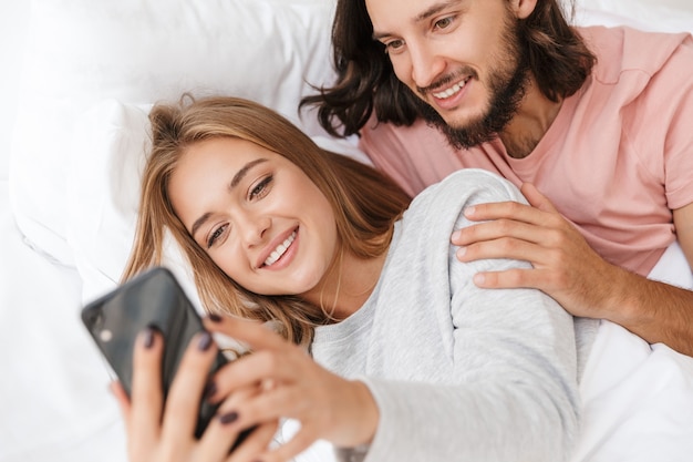 Piękna młoda para leży w łóżku w domu, patrząc na telefon komórkowy
