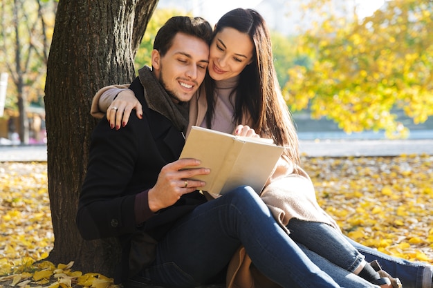Piękna młoda para zakochanych spędzających razem czas w parku jesienią, czytając książkę, siedząc pod drzewem