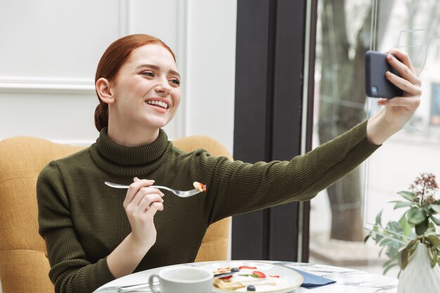 Piękna Młoda Ruda Kobieta Relaksuje Przy Stoliku Kawiarni W Pomieszczeniu, Jedząc Lunch, Biorąc Selfie