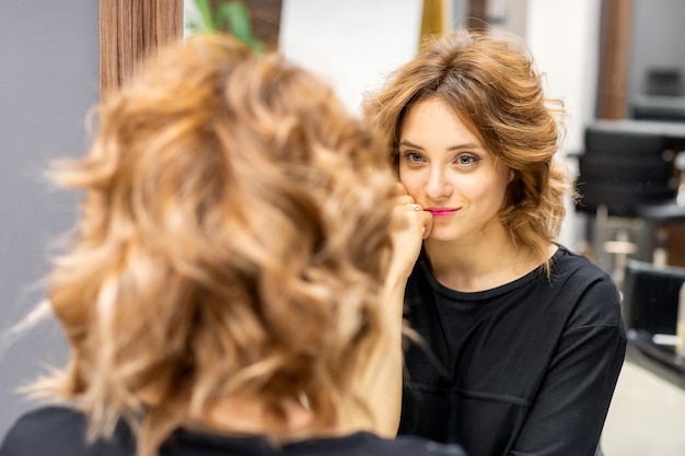 Piękna młoda rudowłosa kaukaska kobieta z nową krótką falującą fryzurą patrząca w lustro sprawdzająca fryzurę i makijaż w salonie fryzjerskim