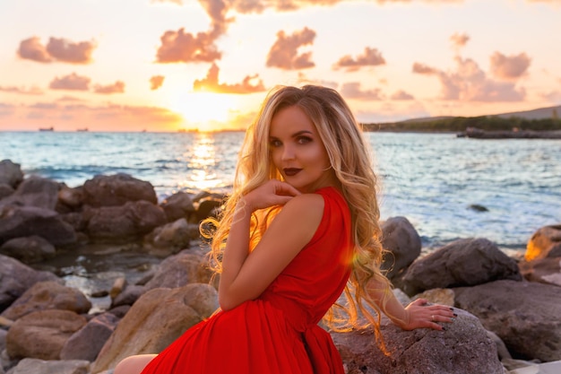 Piękna młoda seksowna blondynka pozuje w wieczorowej czerwonej sukience na nabrzeżu na zachód słońca