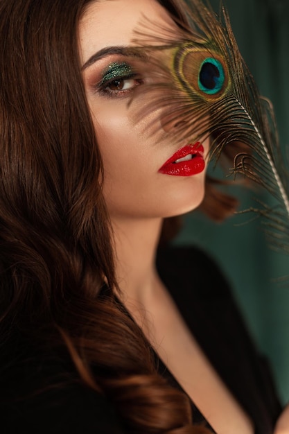 Zdjęcie piękna młoda seksowna modelka z makijażem i fryzurą w czarnym eleganckim garniturze zakrywa oczy kolorowym pawim piórem