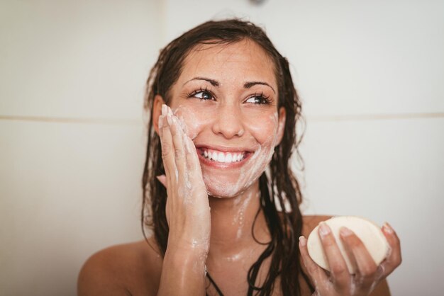 Piękna młoda uśmiechnięta kobieta mycie twarzy mydłem w łazience.