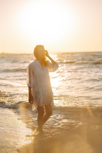 Piękna modelka w eleganckiej białej sukience przy zachodzie słońca z kieliszkiem wina dziewczyna patrzy na wschód słońca