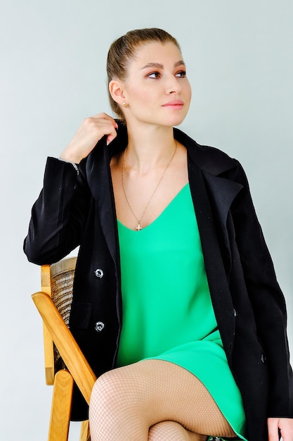 Piękna modna dziewczyna w zielonej sukience i czarnej kurtce pozuje siedząc na krześle na szarym tle