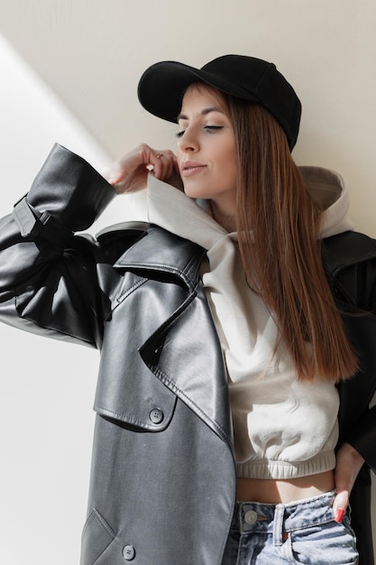 Zdjęcie piękna modna młoda hipster kobieta z modną czarną czapką w stylowym skórzanym płaszczu z modną bluzą z kapturem i stoi w pobliżu beżowej ściany w słońcu miejski styl kobiecy