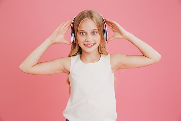 Piękna nastolatka 8-10 w ubranie, słuchanie muzyki i dotykanie słuchawek bezprzewodowych na głowie, odizolowane na różowym tle