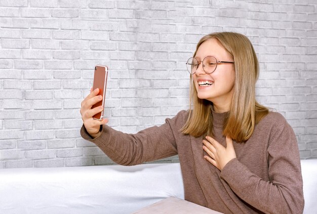 Piękna nastolatka w okularach patrzy na telefon i śmieje się. Dziewczyna śmieje się podczas rozmowy wideo. Koncepcja komunikacji nastolatków. Komunikacja z rodziną i przyjaciółmi.