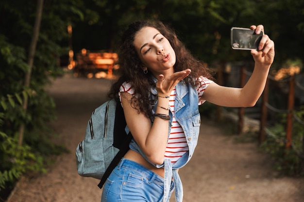 Piękna Nastolatka Z Plecakiem, Dmuchanie Buziaka Na Aparat Telefonu Komórkowego I Robienie Selfie Podczas Spaceru Na świeżym Powietrzu