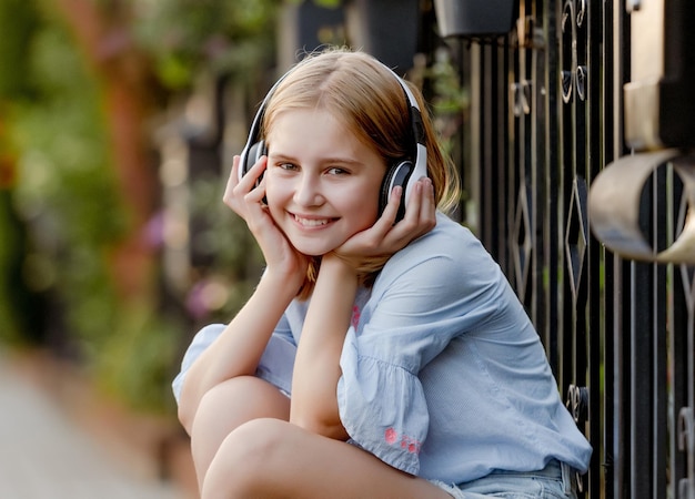 Zdjęcie piękna nastolatka z słuchawkami siedząca w pobliżu ogrodzenia na ulicy i uśmiechająca się uroczo.