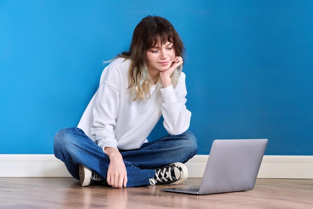 Piękna nastoletnia kobieta siedzi na podłodze z laptopem na niebieskim tle