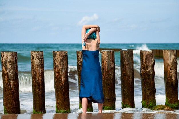 Piękna niebieskowłosa kobieta performer w ciemnoniebieskiej sukience stojącej na plaży, trzymając pędzel, widok z tyłu. Współczesna sztuka abstrakcyjna, sztuka tańca w przyrodzie, nowoczesna sztuka performance na świeżym powietrzu