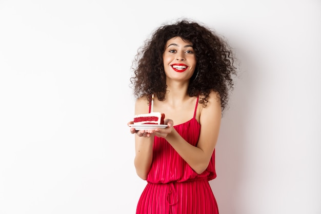 Piękna pani w czerwonej sukience obchodzi urodziny, trzymając kawałek ciasta ze świecą i uśmiechnięty, stojący szczęśliwy na białym tle.