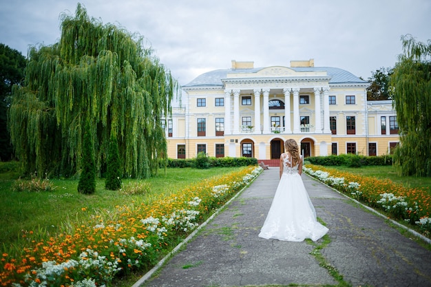Piękna panna młoda w białej sukni z trenem i spacer na tle dużego domu z kolumnami w dniu ślubu