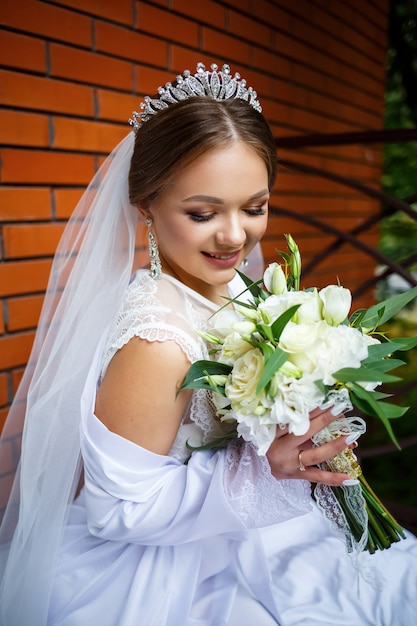 Piękna panna młoda w welonie i białym płaszczu siedzi na kocu z bukietem ślubnym w dłoniach