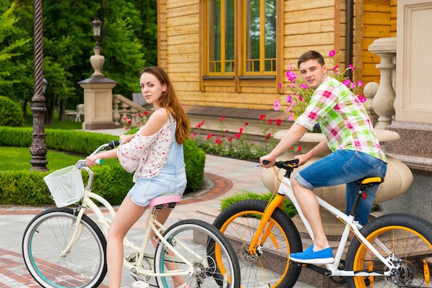 Piękna para odwróciła się i spojrzała w obiektyw podczas jazdy na rowerze na tle luksusowego domu w parku