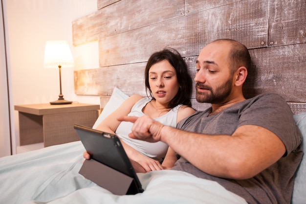 Piękna para sobie piżamę, leżąc w łóżku, surfując po internecie przy użyciu komputera typu tablet.