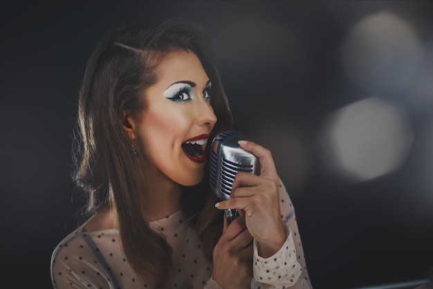 Piękna piosenkarka ze stylowymi włosami i makijażem w stylu retro trzyma vintage mikrofon ze statywem i śpiewa.