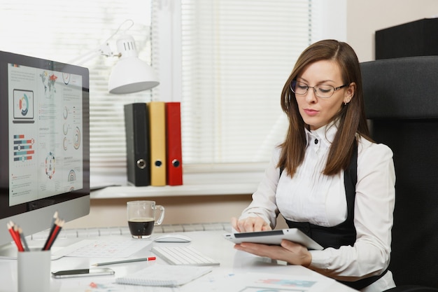 Piękna pochłonięta biznesowa kobieta o brązowych włosach w garniturze i okularach siedząca przy biurku z filiżanką kawy, telefonem komórkowym, pracująca przy komputerze z nowoczesnym monitorem z dokumentem i tabletem w jasnym biurze