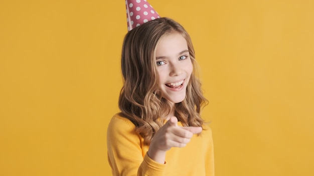 Piękna podekscytowana blond nastolatka w kapeluszu urodzinowym, wyglądająca wesoło, wskazując palcem wskazującym na aparacie na żółtym tle