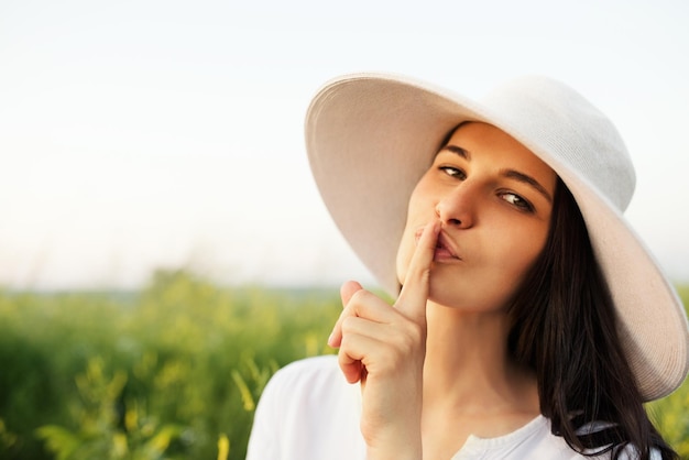 Piękna romantyczna młoda kobieta w kapeluszu i białej koszuli milczy z palcem na ustach na tle pola przyrody
