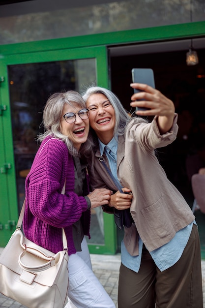 Piękna roześmiana dojrzała azjatycka dama z towarzyszem robi selfie ze smartfonem na ulicy miasta