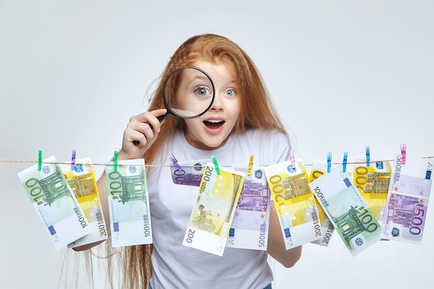 Piękna rudowłosa dziewczyna bada banknoty euro przez lupę.