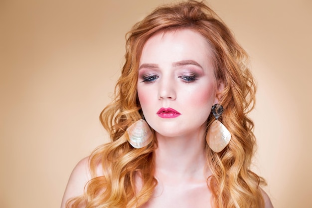 Piękna rudowłosa dziewczyna z falującymi włosami i makijażem pozuje na beżowym tle w studiu w dużych złotych kolczykach