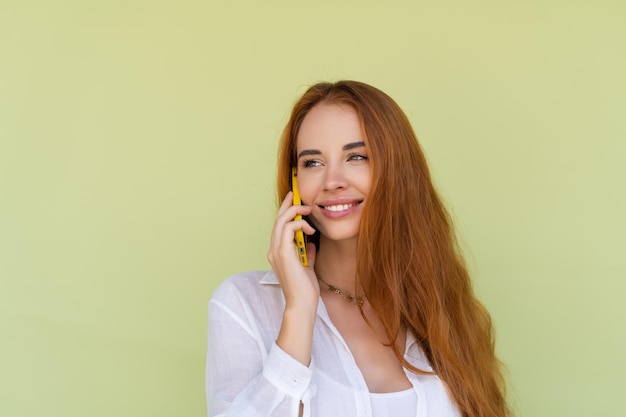 Piękna rudowłosa kobieta w swobodnej koszuli na zielonym tle rozmawia przez telefon komórkowy, uśmiechając się i śmiejąc