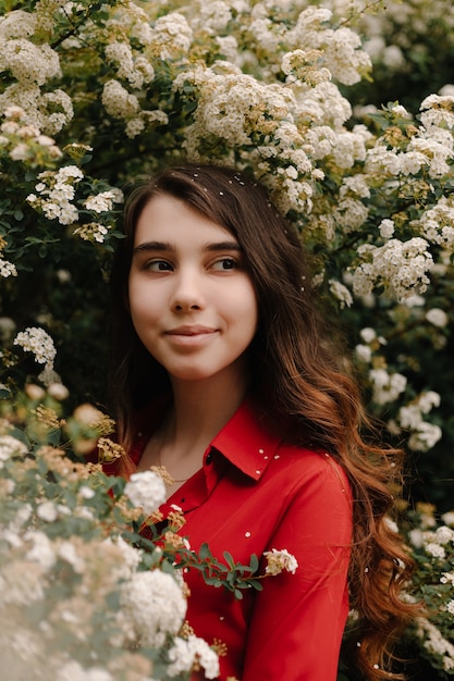 Piękna rudowłosa młoda dziewczyna w czerwonej bluzce na wiosnę w pobliżu kwitnących krzewów