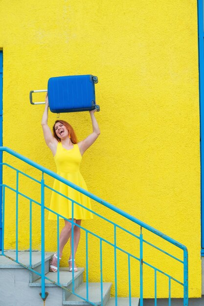 Piękna rudowłosa uśmiechnięta kobieta radośnie schodzi po schodach i macha niebieską walizką na żółtym tle