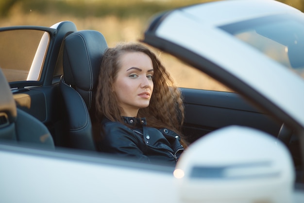 Piękna seksowna dziewczyna z długie włosy w skórzanej kurtce i rzemiennych spodniach w okularach przeciwsłonecznych siedzi w drogim samochodzie przy zmierzchem.