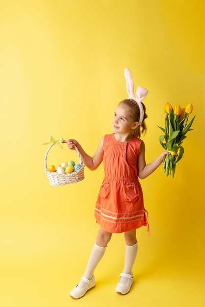 Piękna śliczna mała dziewczynka w uszach zajączka wielkanocnego trzyma kosz z jajkami i wiosennym bukietem