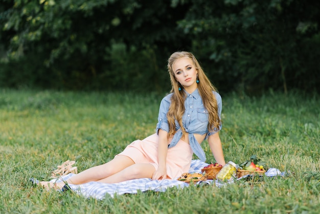 Piękna słodka dziewczyna w niebieskiej dżinsowej koszuli i szyfonowej różowej spódnicy siedzi na szkockiej kracie na letnim pikniku
