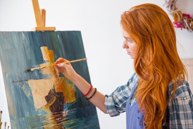 Piękna spokojna młoda malarka z długimi rudymi włosami maluje obraz na płótnie w warsztacie artystycznym