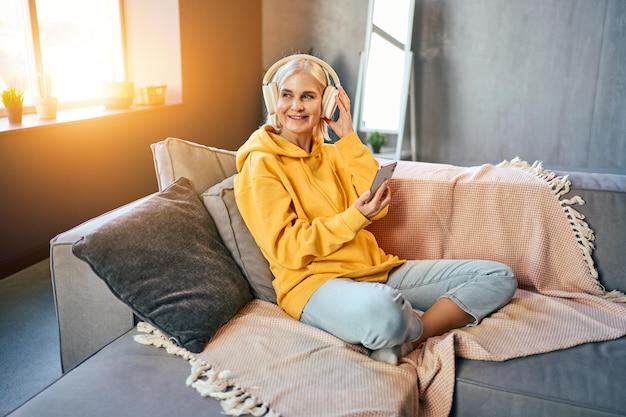 Piękna starsza kobieta siedzi na sofie w żółtych dżinsach z kapturem i słucha muzyki w bezprzewodowych słuchawkach z telefonem w dłoniach i odwraca wzrok