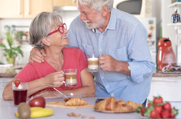 Piękna starsza para uśmiecha się śniadanie w domu. Szczęśliwi emeryci pijący cappuccino jedzący owoce i rogalika
