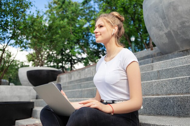 piękna stylowa młoda dziewczyna w słuchawkach bezprzewodowych na sobie białą koszulkę z laptopem na zewnątrz