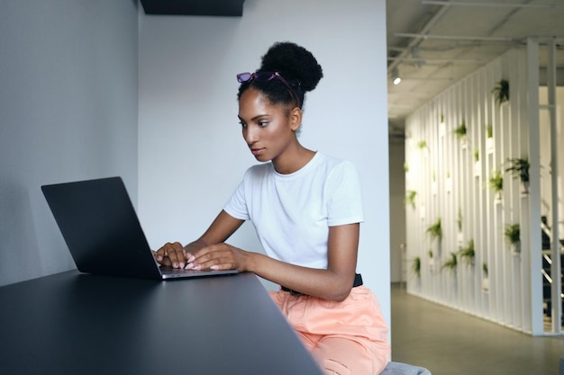 Piękna, swobodna, afroamerykańska dziewczyna pewnie pracująca na laptopie w nowoczesnej przestrzeni coworkingowej