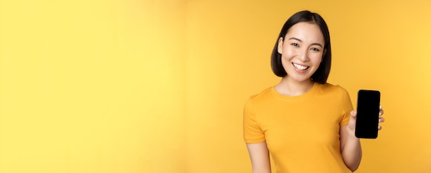 Piękna szczęśliwa azjatycka dziewczyna pokazująca aplikację ekranu telefonu komórkowego na gadżecie smartfona stojącego na żółtym tle