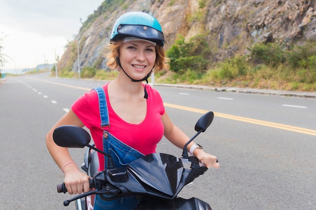 Piękna Szczęśliwa Dziewczyna, Młoda Kobieta, Rowerzysta Lub Motocyklista Jedzie Motocyklem, Motorowerem Lub Rowerem, Uśmiechając Się. żeński Jeździec W Hełmie Dla Zbawczej Jazdy Na Drodze W Górach W Letnim Dniu.