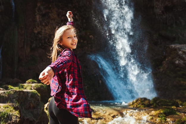 Piękna szczęśliwa dziewczynka cieszy się krajobrazem w górach, wznosząc ręce i patrząc na piękny wodospad na świeżym powietrzu. Jej włosy rozwiewa wiatr.